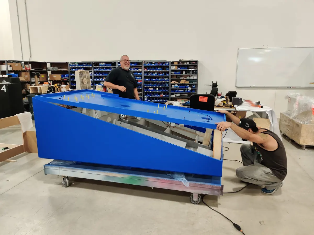 Two people test-fitting the blue playfield cabinet on a rolling frame in the shop