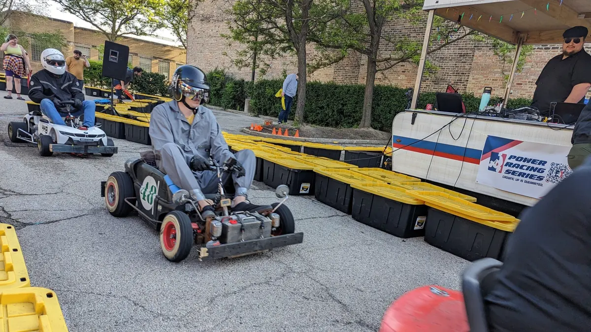 A driver in a helmet piloting a modified ride-on kart past the timing tent