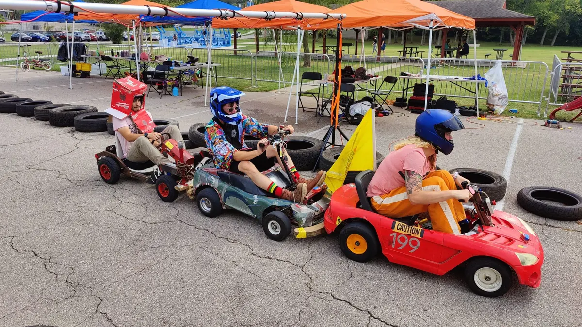 Three helmeted drivers racing modified children’s ride-on cars around a tire-lined parking-lot track