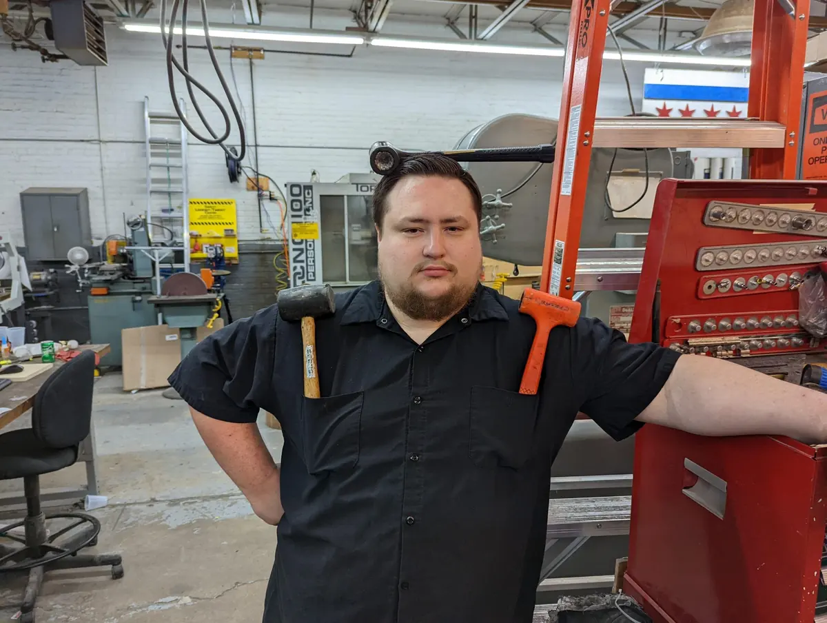 James holding a hammer in each hand beside a red tool chest in the shop