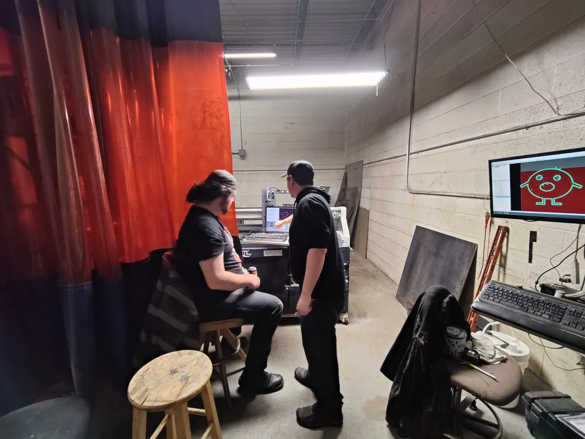Two members at the CNC plasma cutter’s control station behind an orange curtain
